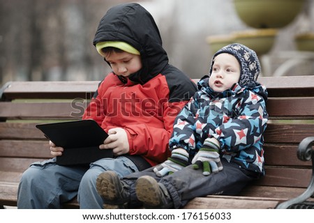 Young brothers sit together on a wooden park bench in warm winter clothing with the older child using a tablet computer
