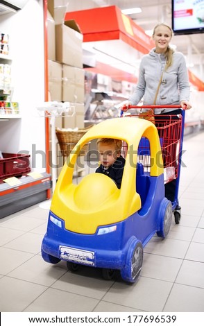 Child friendly supermarket shopping with a young mother pushing her small son in a colourful plastic car attached to the shopping trolley keeping him happy and entertained while she buys the groceries