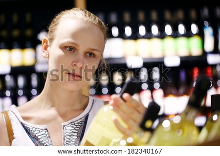 Woman shopping for wine or other alcohol in a bottle store standing in front of shelves full of bottles with a serious expression as she tries to make up her mind