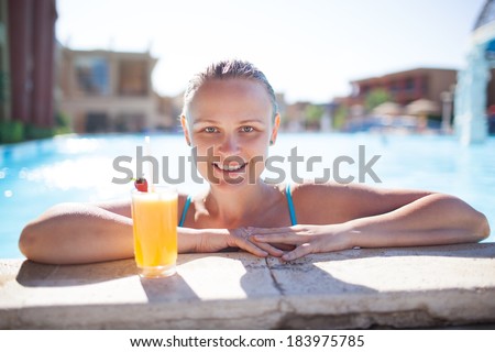 Smiling young woman enjoying a refreshing orange drink or cocktail in the pool resting her arms on the pool surround smiling at the camera