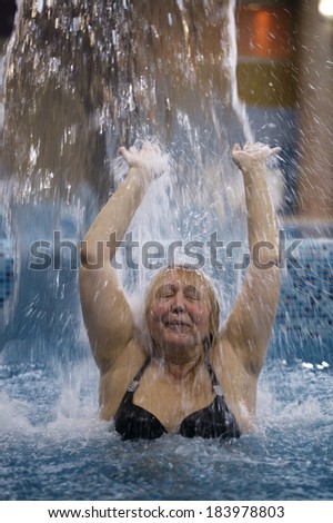 Middle-aged woman splashing in a pool under a jet of water with her arms raised and eyes closed in enjoyment