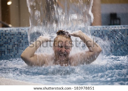 Overweight middle-aged woman playing under a water fountain splashing into a swimming pool as she stands chest deep in water laughing as the cascade pours over her head