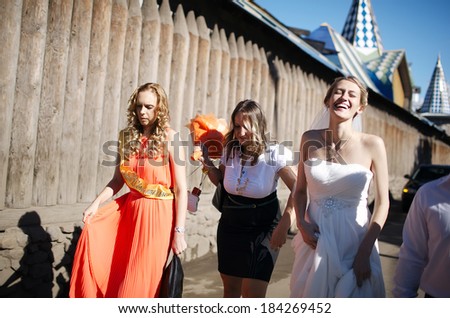 Laughing happy bride and her bridesmaid walking with their friends along a road outdoors alongside a wooden palisade