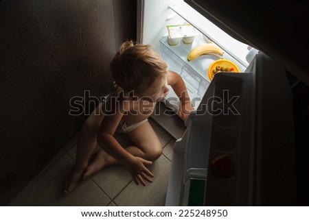 Little hungry boy sitting on the floor by open fridge at night. He searching something tasty to eat