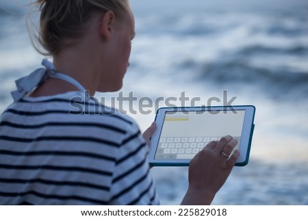 Young woman typing on touchpad sitting alone on the beach. Blurry sea in background