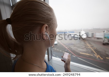 Woman in headphones by the window at aiport listening to music using smart phone. Entertainment during flight waiting