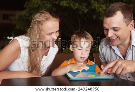 Young parents and little son in a cafe in the evening. Father and son using touch pad, mother watching them