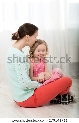 Young mother and beautiful little daughter sitting on the floor, mom kissing little girls head