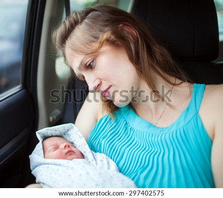Young mother holding newborn wrapped in baby blanket while sitting in the car. Woman looking at her baby with deep feelings expressed in the eyes