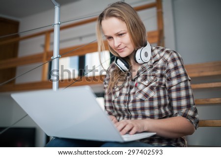 Pretty young woman with headphones using laptop for chatting or entertainment sitting on the stairs at home