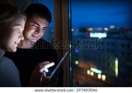 Two friends are standing by window with tablet PC. Backlit screen is lighting their faces.