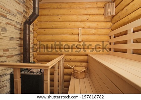Inside view in traditional Russian log sauna with wood-burning heater, wooden benches, lamps and sandglass on the walls and bucket with spoon on bench