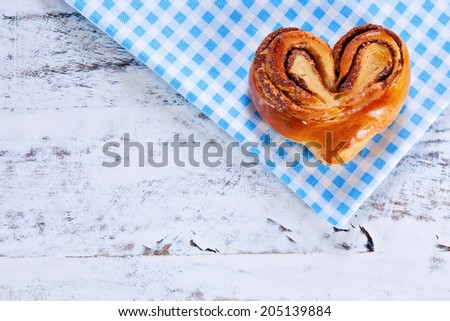Rustic home-made heart shape freshly backed cinnamon roll on blue checkered cloth over white painted board or table. Above view