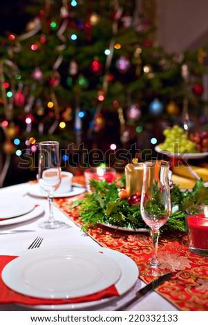 Festive dinner table setting with red table cloth, champagne glasses, candles and Christmas tree on the background