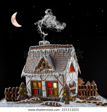 Decorative gingerbread house with lights inside and smoke coming out the chimney on black background with moon and stars . Rural Christmas night scene