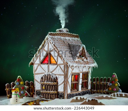 Decorative gingerbread house with lights inside and smoke coming out the chimney with Northern lights on background. Rural Christmas night scene