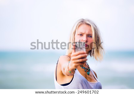Beautiful young blonde blue eyes woman taking a selfie on smart-phone outdoor at the rocky sea shore. Trendy fashion female model dressed in white top