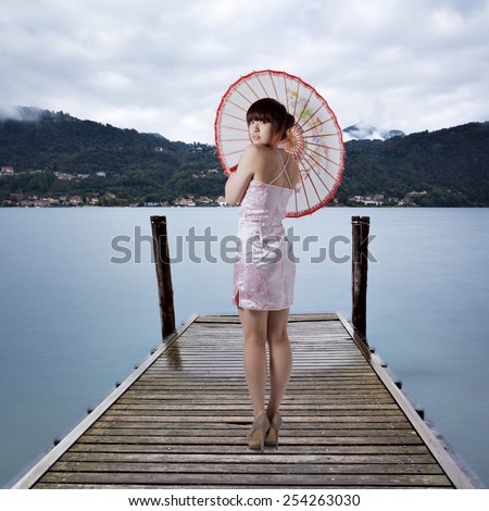 Young pretty chinese girl dressed in traditional pink national dress posing on old wooden pier with Pella town and misty Italian Alps mountains on background. Isola San Giulio on Lago d\'Orta