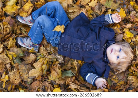 Small blond boy with blue eyes lays on bed of autumn fallen leaves