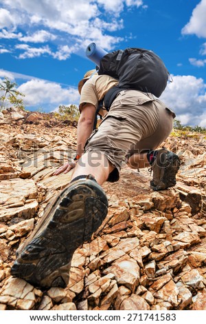 Close up of a man climbing up red rock dressed in hiking shoes, khaki color shorts and t-shirt with black rucksack.