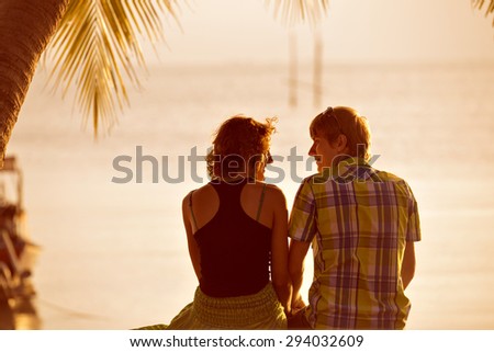 Young couple sit together holding hands under a palm tree and looking at each other. Lovers on vacation