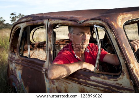 Old car, man in his 50's behind the wheel of a rusted out, vintage type car in an Australian field.