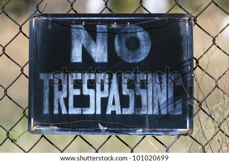 Close up of a no trespassing sign attached to a chain link fence. The sign is black and white and well aged, as is the chain link fence./ No Trespassing, Period! / This means you.