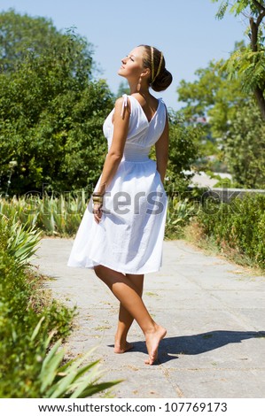 Alluring brown-haired woman outdoors in Greek goddess style in luxury dress standing back. Stylish romantic girl in a white sundress walking in the park. romantic concept. Amazing Nymph. Greece