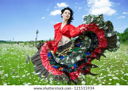 beautiful gypsy woman dancing in a green field of daisies. attractive dark-haired woman dressed like a gypsy on the background of a green field and blue sky