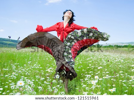beautiful gypsy woman with head thrown back dancing in a green field of daisies. attractive dark-haired woman dressed like a gypsy on the background of a green field and blue sky