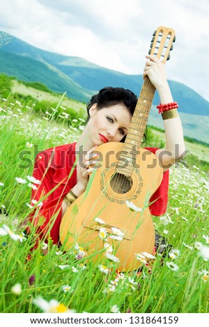 illustration of alluring gypsy woman holding guitar in hands and sitting in a green field of daisies.dark-haired woman dressed like a gypsy with a guitar on a background of green field, hills and sky