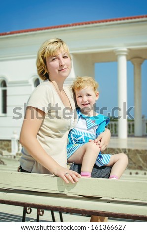 happy mother and adorable baby daughter posing outdoors. smiling family. beautiful blond woman and funny little girl in trendy wear. mother and daughter sitting on the wooden bench in summer park