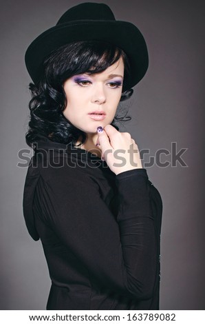 Beautiful brunette woman in stylish black hat posing in studio. Romantic lady in black on gray background. Fashion portrait.