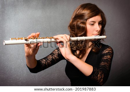 Beautiful young woman playing flute in studio. Alluring flutist in black evening dress with hairstyle makeup posing with flute on gray background