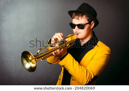 Handsome jazz trumpet player in hat, yellow jacket and black glasses playing trumpet. Portrait of young musician playing the trumpet at studio.