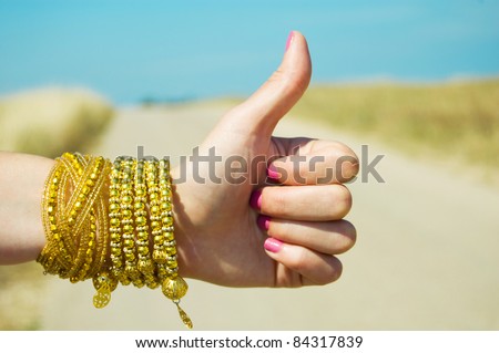 hitchhiking, thumbs up female hand on the background of the road and blue sky, for praise or like hand gestures. female hand with pink manicure and yellow gold bracelets