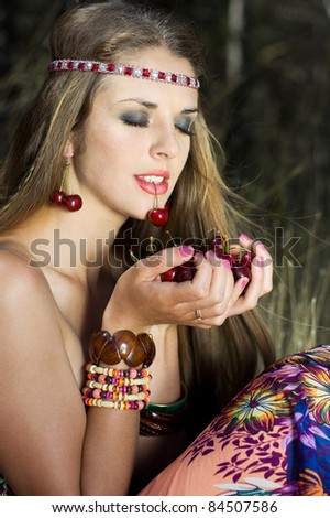 alluring hippie woman holding a cherry in her hands. resting attractive long-haired woman with closed eyes with earrings in the form of cherries eating cherry sitting in the forest outdoors