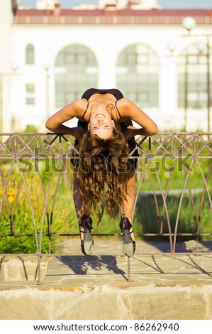 alluring redheaded smiling woman lying upside down on a forged fence. Stylish slim tanned girl with healthy curly hair on the background of green lawn and building with large windows. outdoors in park
