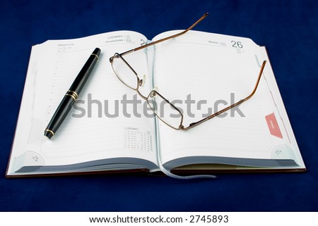 A opened timetable with a pen with a gilded clip and steel-rimmed glasses on it.