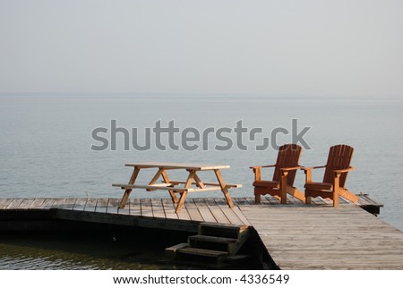 Two adirondack wooden chairs on dock facing a blue lake with reflections