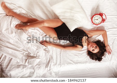 Sexy lazy girl in black body lying with red alarm clock on the bed. Young attractive woman relaxing lazing in her bedroom at the morning.