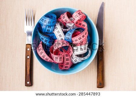 Diet food healthy lifestyle and slim body concept. Many colorful measuring tapes in blue bowl on table with knife and fork, top view
