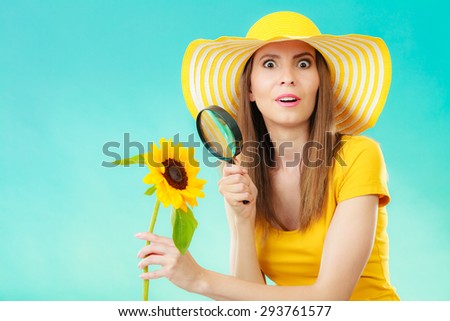 Botanist woman surprised face expression in yellow hat examining flower looking through magnifying glass on blue background