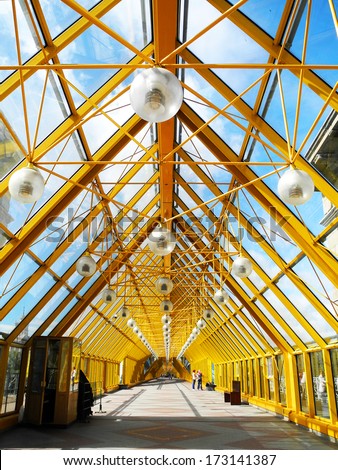 Yellow bridge of trusses and beams. Indoor glass bridge for pedestrians. Sunny day, bright light and contrasting shadows. Path, road, lights
