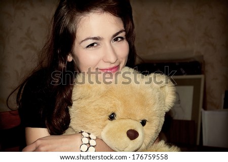 Color photo. Portrait of c girl with dark hair and brown eyes. Smile, a kind face. Beige teddy bear, children's toy, bow in the box. White bracelet on his arm, costume jewelery