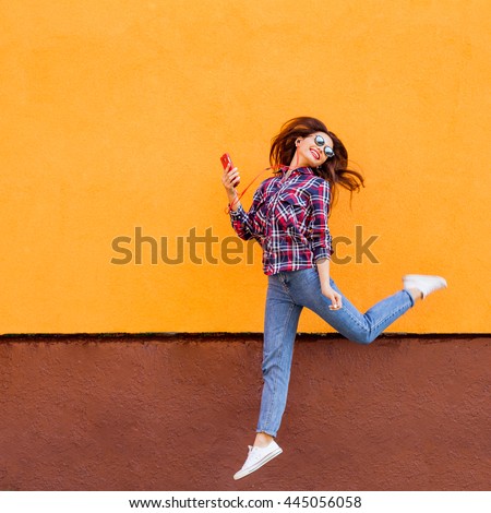Fashion portrait of pretty smiling and jumping woman in sunglasses with smartphone against the colorful orange wall. Copyspace