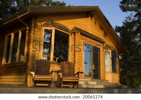 A cozy log cabin in the woods is fronted by a pair of wooden chairs.