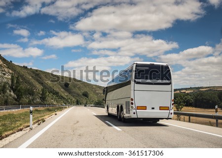 A white coach, or long haul bus for tourists drives through the open roads of Spain, Europe on a summer day. There are white clouds against a blue sky and countryside.