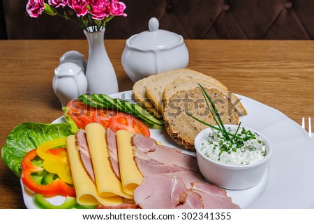 Small bowl of cottage cheese on a plate with slices of home-smoked tenderloin, cheese, smoked ham, tomato, cucumber and Polish bread.