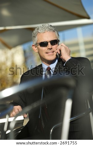 Prime adult Caucasian man in suit sitting at ouside patio table wearing sunglasses and talking on cellphone smiling.
